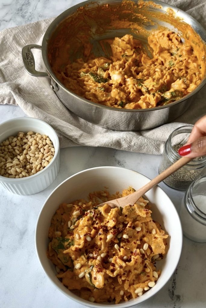 A bowl of creamy red pepper pasta garnished with pine nuts and red pepper flakes.